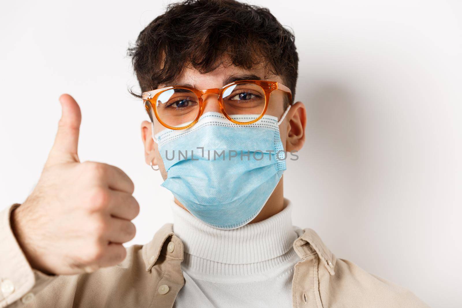 Portrait of young man in glasses and medical mask showing thumb up, smiling with eyes, standing on white background. Covid-19 and pandemic concept by Benzoix