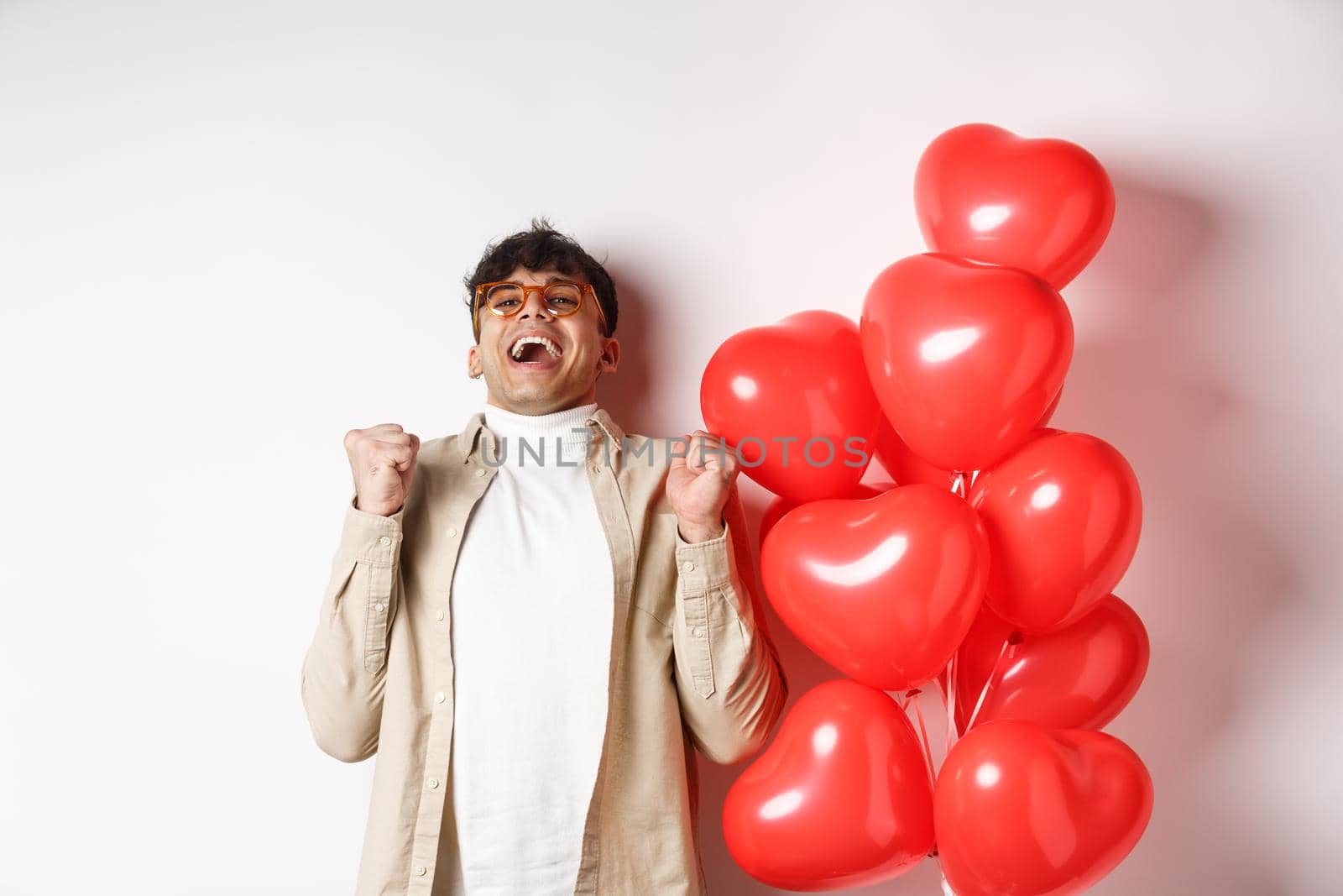 Valentines day. Happy modern man celebrating, screaming from joy and happiness, having date with lover, being in love, standing near heart balloons on white background.