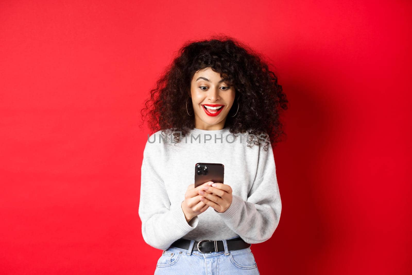 Image of young woman with curly hair, reading message on smartphone and smiling happy, receiving good news online, standing on red background.