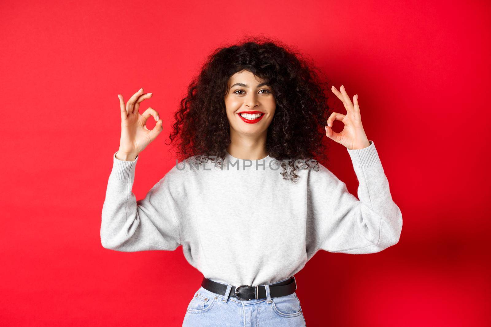 Confident pretty girl with curly hairstyle, showing okay gestures and smiling, approve and agree with you, praising excellent choice, standing satisfied on red background by Benzoix