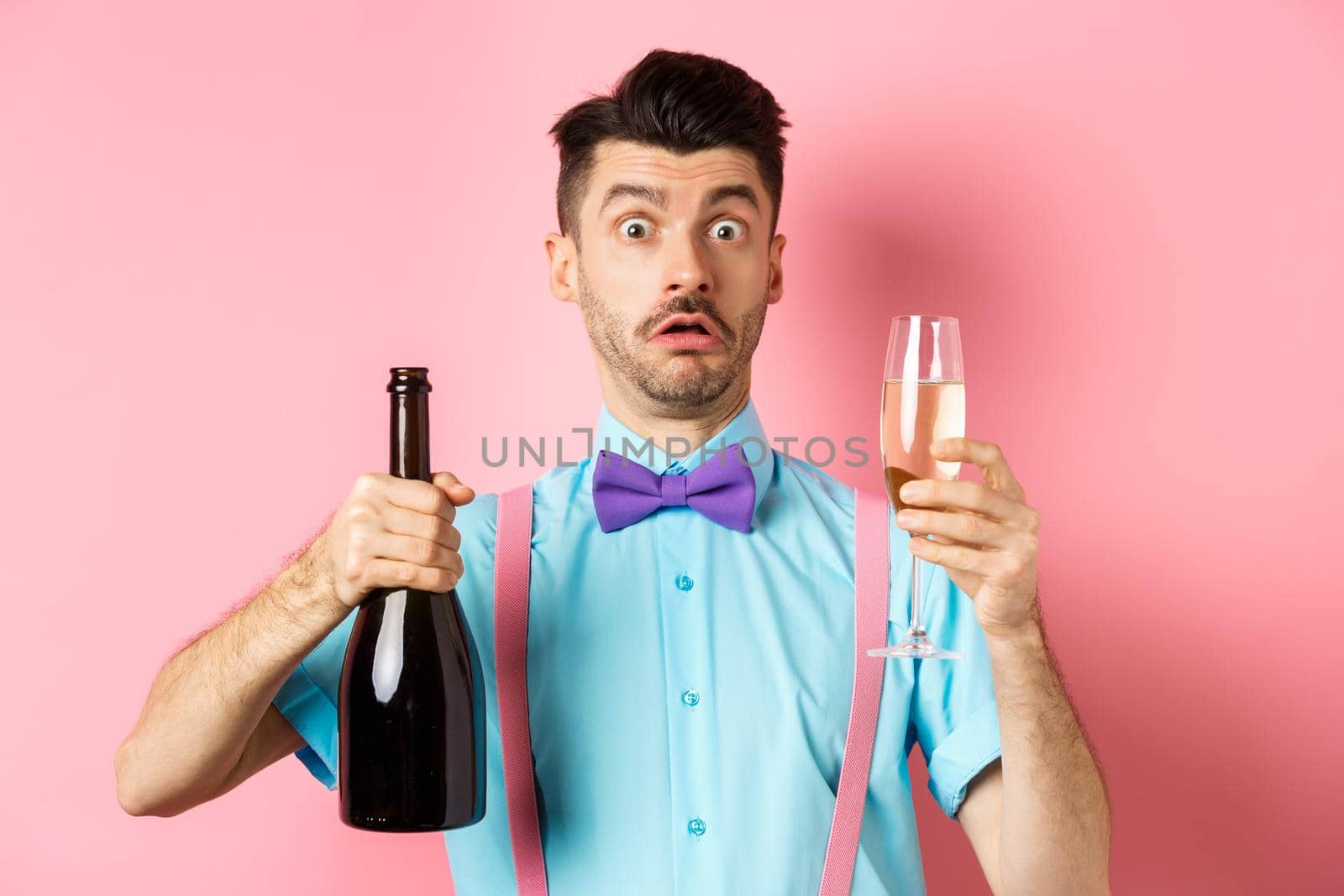 Holidays and celebration concept. Confused man staring startled at camera while pouring glass of champagne, standing puzzled over pink background.
