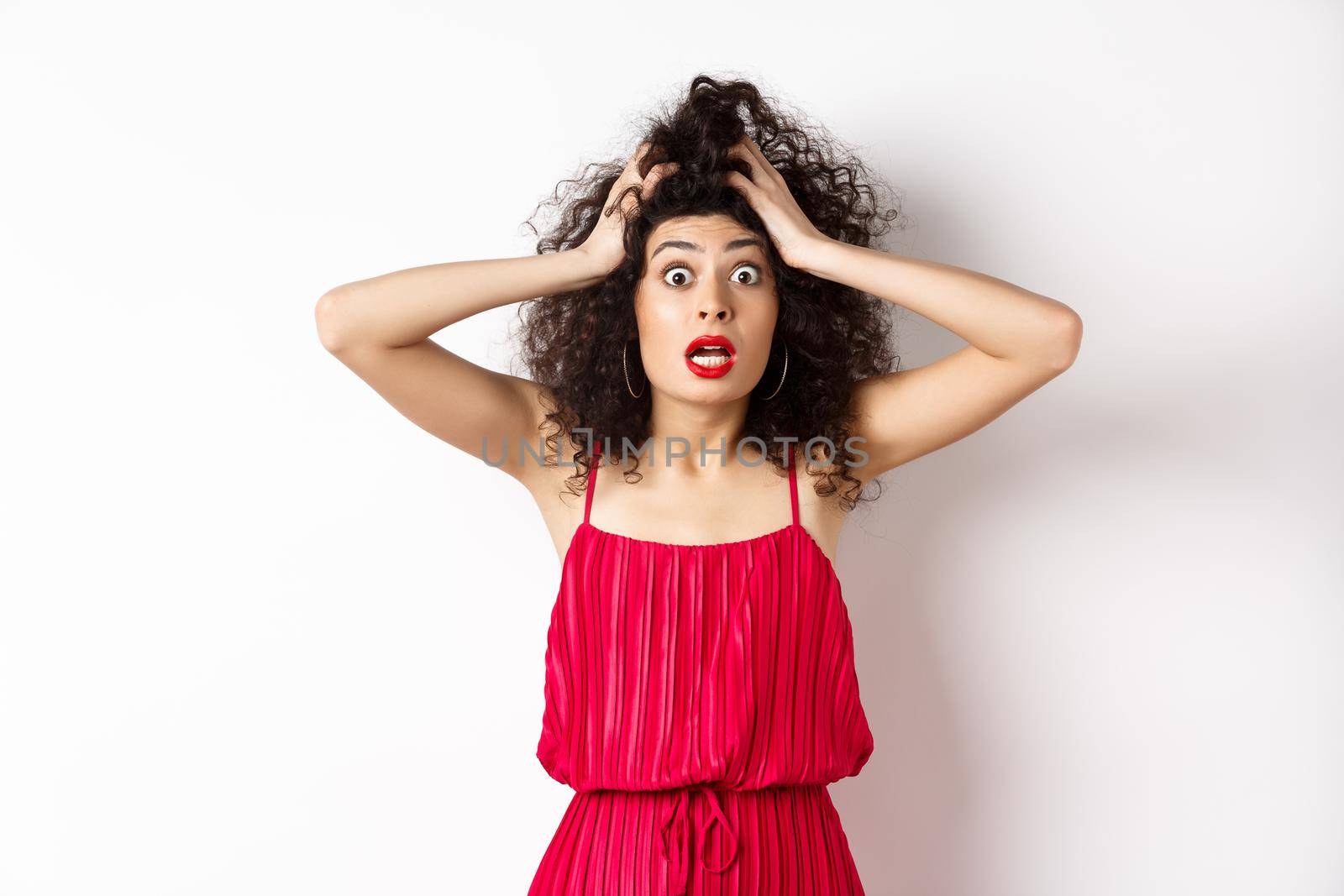 Shocked young woman holding hands on head and panicking, staring frustrated at camera, wearing red dress and makeup, white background.