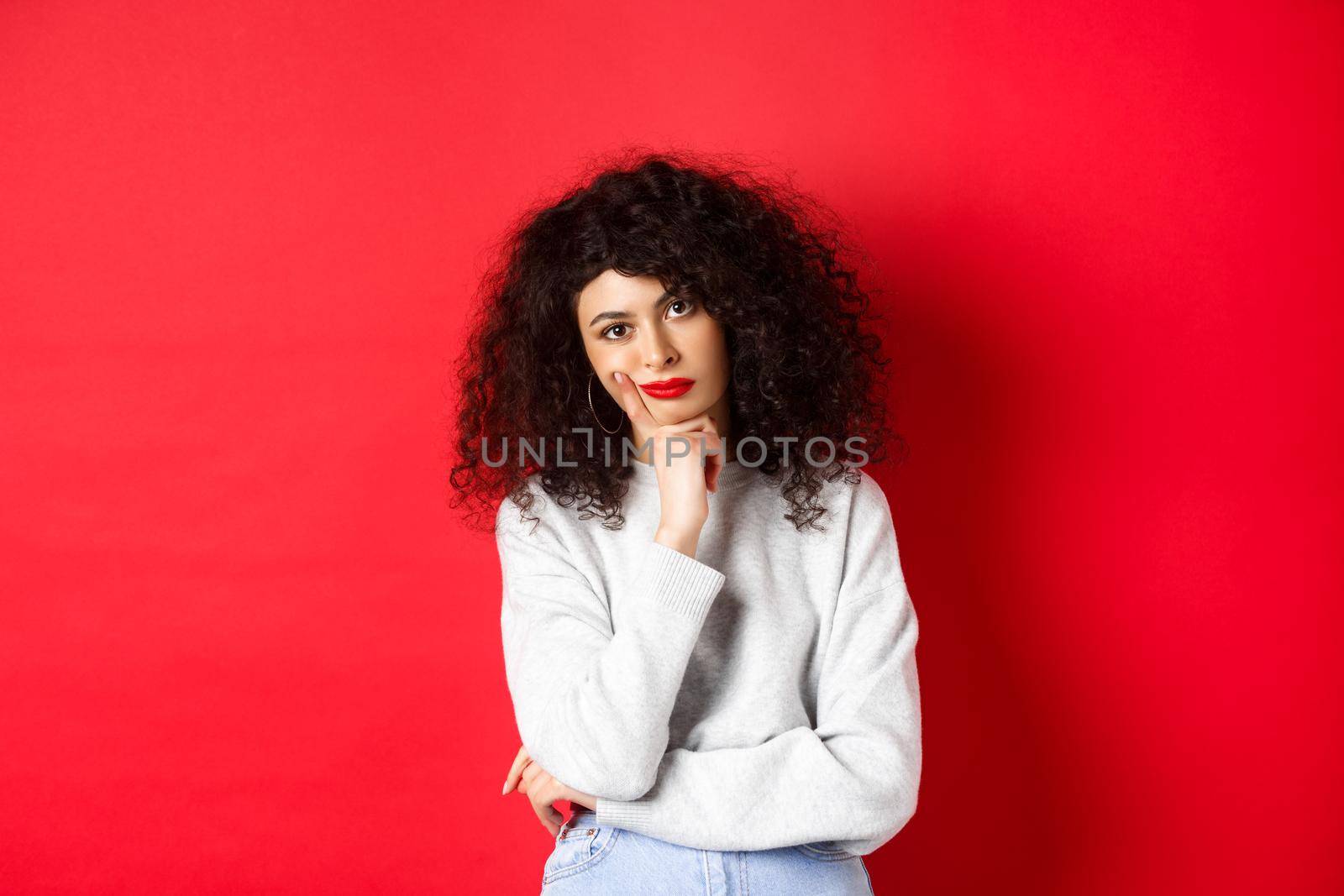 Bored and tired curly woman with red lips, looking bothered at camera, standing against studio background.