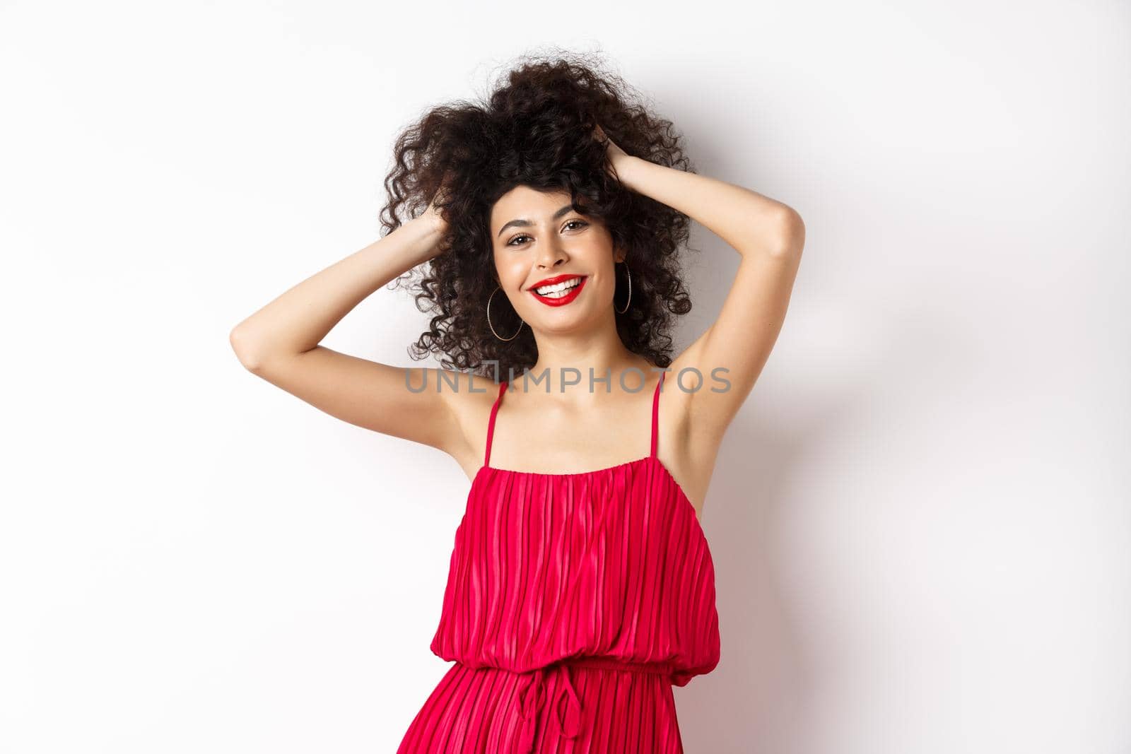 Happy smiling woman touching her curly hair and laughing, standing in red dress on white background.