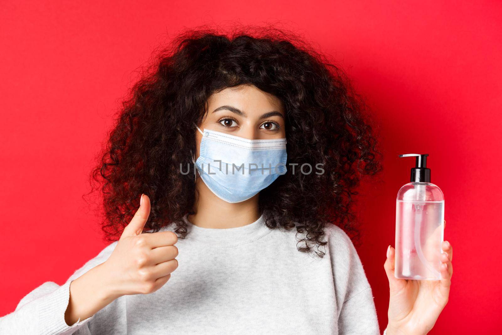 Covid-19, social distancing and quarantine concept. Portrait of woman in face mask from coronavirus, showing hand sanitizer bottle and thumb up, recommending antiseptic.