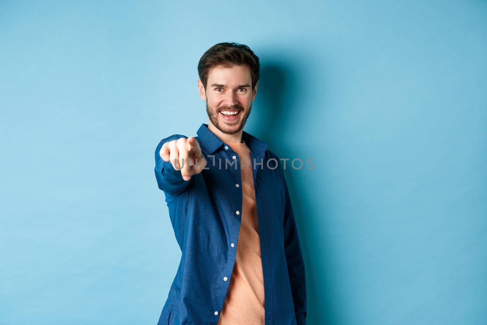Confident smiling guy choosing or inviting you, pointing finger at camera decisive, standing on blue background. Copy space
