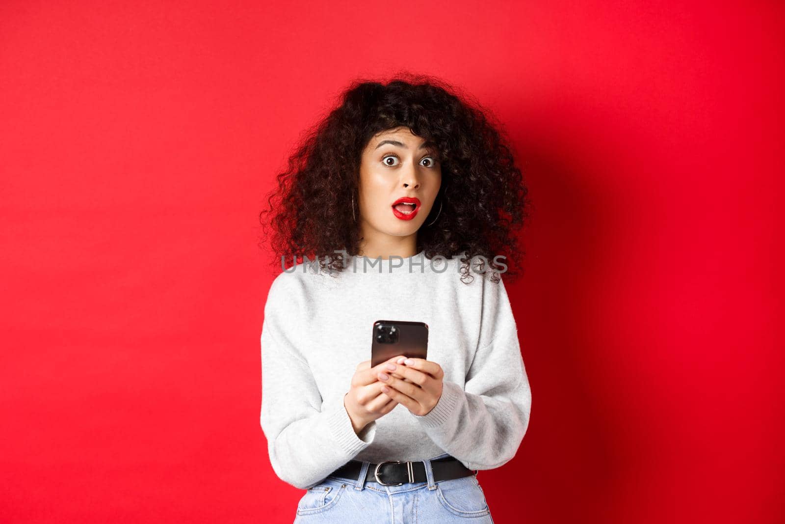 Image of pretty curly woman with shocked face, reading message on smartphone startled, standing against red background.