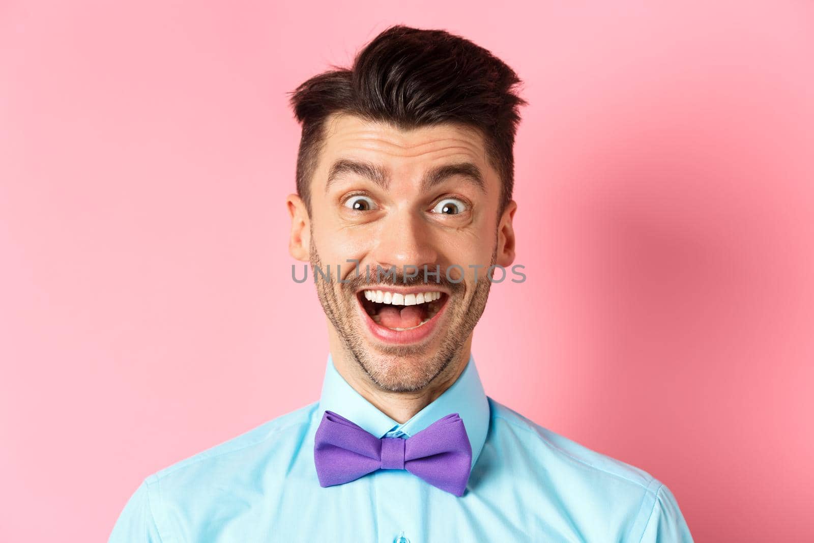 Close-up of surprised and happy young guy, open mouth and smiling amazed, checking out promo offer, standing in bow-tie over pink background.