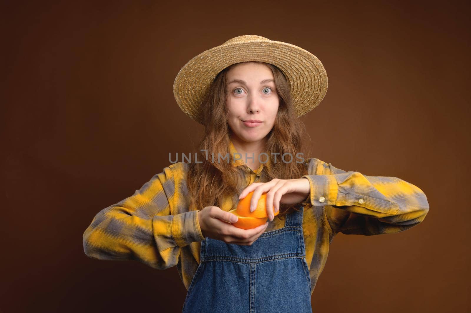 Attractive rustic Caucasian young woman in straw hat holding cut orange fruit. Studio portrait over brown background. Looks into the camera.