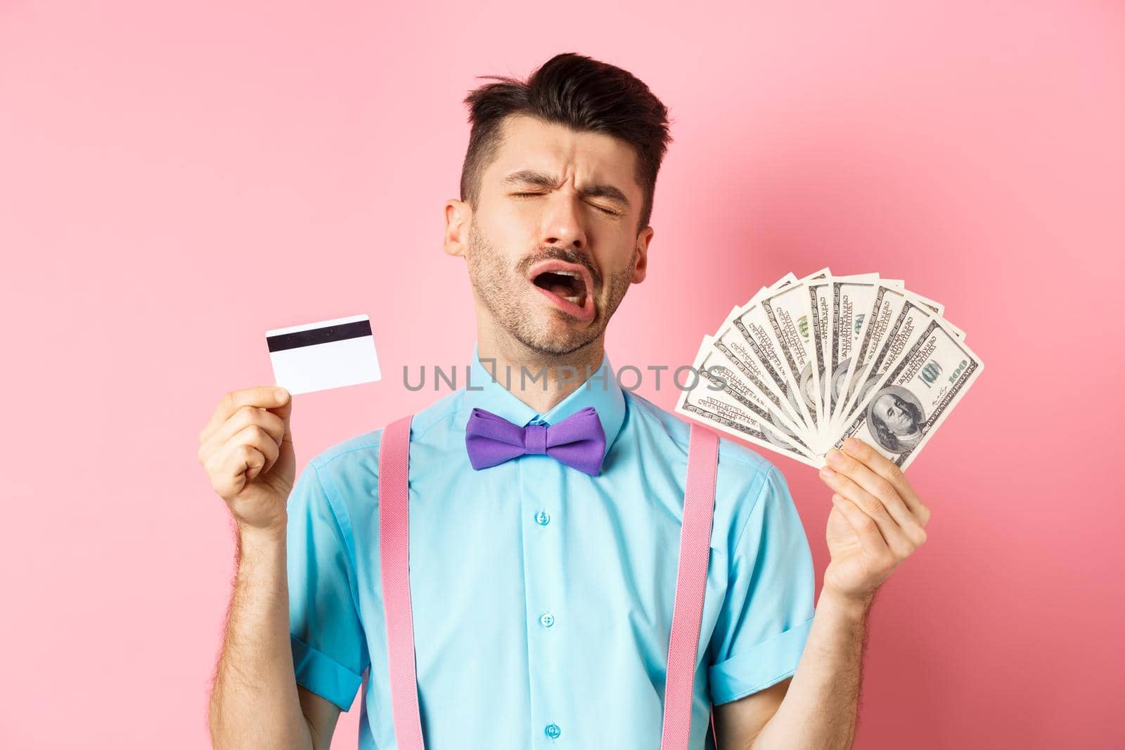 Sad crying man looking miserable, showing plastic credit card and money, standing upset on pink background by Benzoix