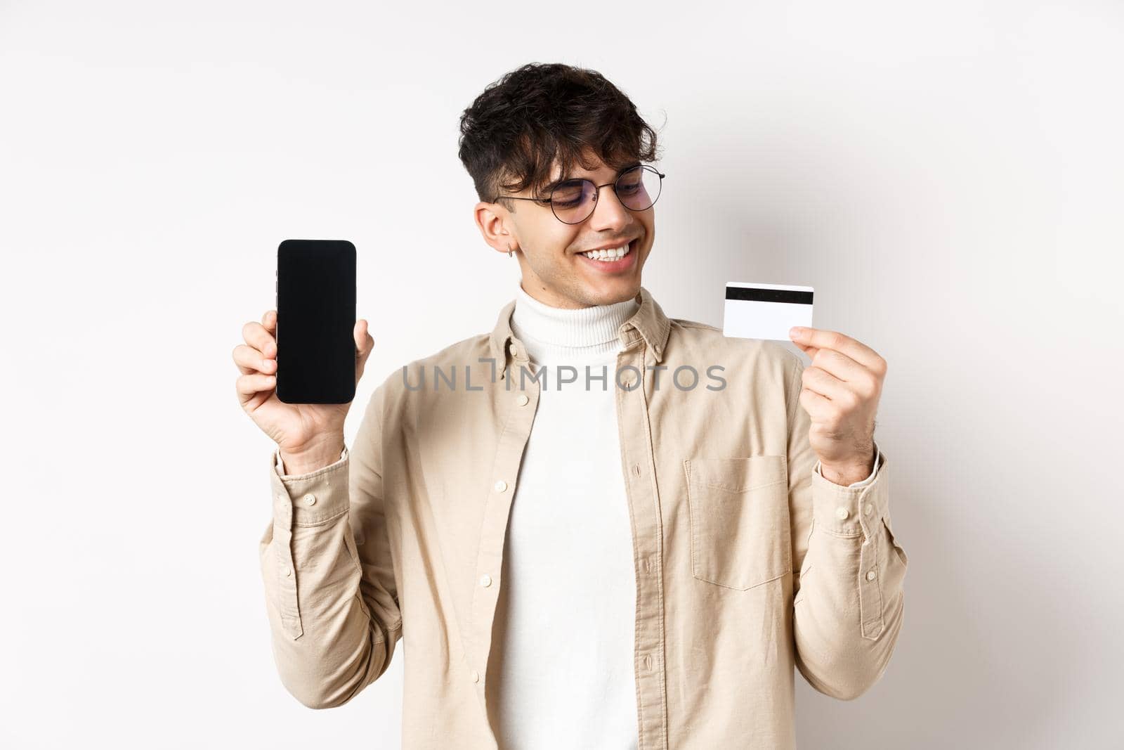Satisfied young man looking pleased at credit card, showing empty smartphone screen, standing on white background.