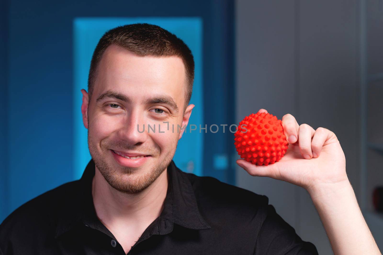 Portrait of a young Caucasian physiotherapist masseur on a blue background with a red massage ball in his hands. Smiling and looking at the camera. Self-massage and self-treatment concept by yanik88