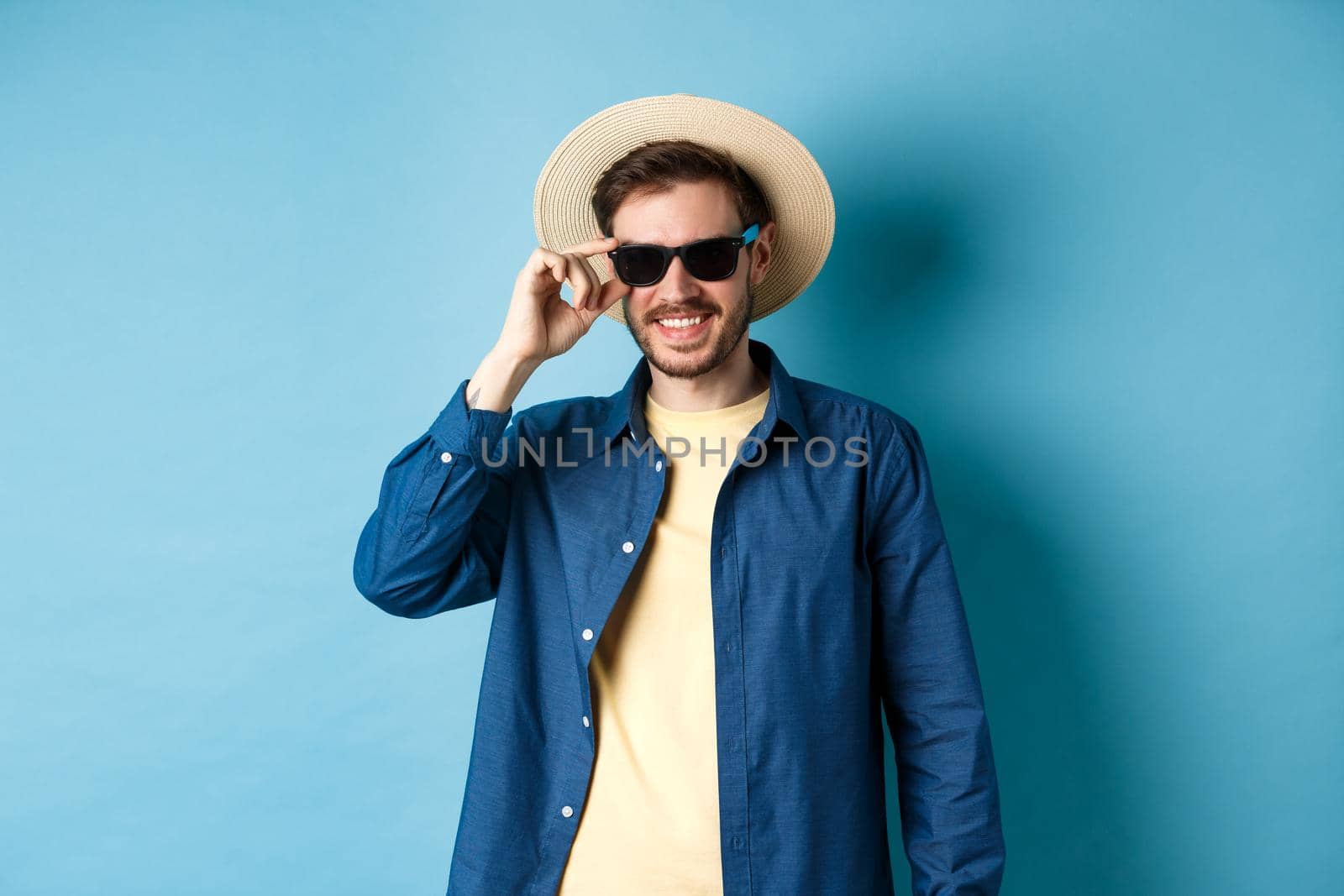 Happy smiling guy going on summer vacation, wearing straw hat and black sunglasses, standing on blue background.