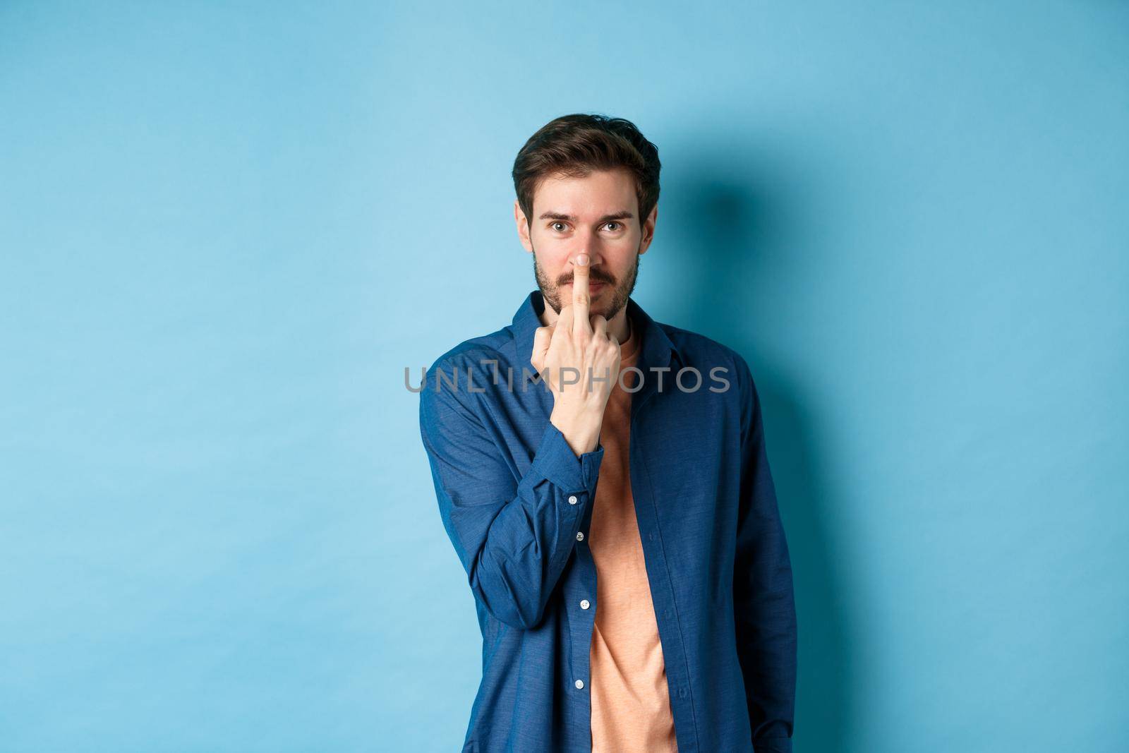 Ignorant and rude guy showing middle finger and smiling, say fuck you, standing on blue background.