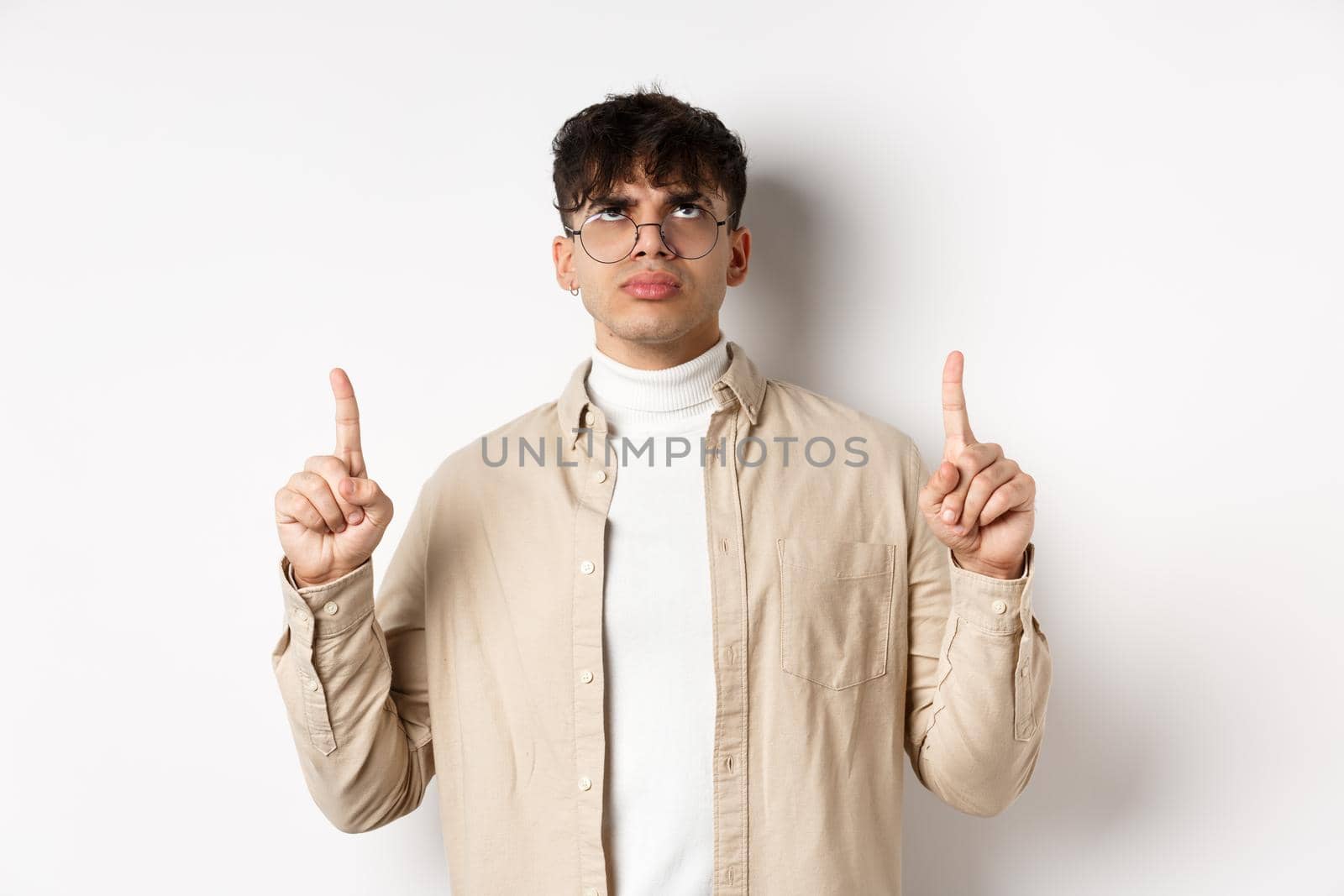 Image of annoyed or offended young man looking, pointing fingers up bothered, frowning disappointed, standing on white background.