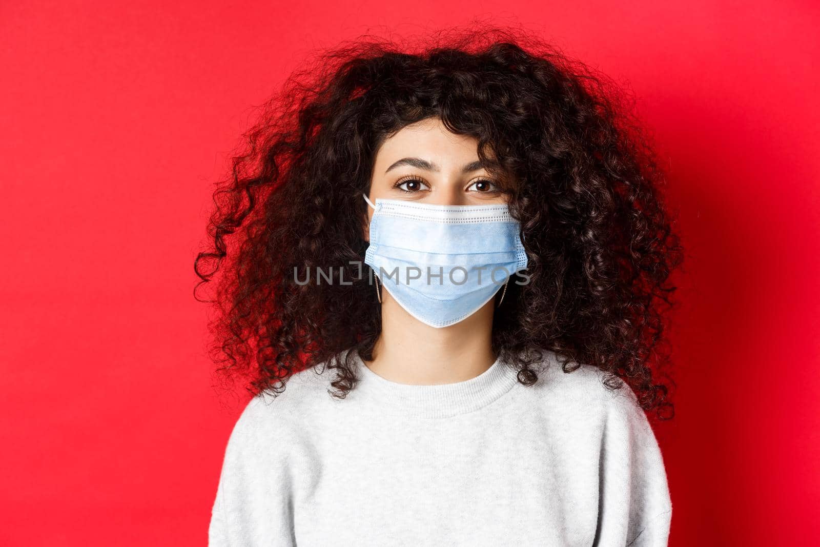 Covid-19 and pandemic concept. Close-up of modern young woman with curly hair, wearing medical mask from coronavirus, smiling at camera, red background by Benzoix