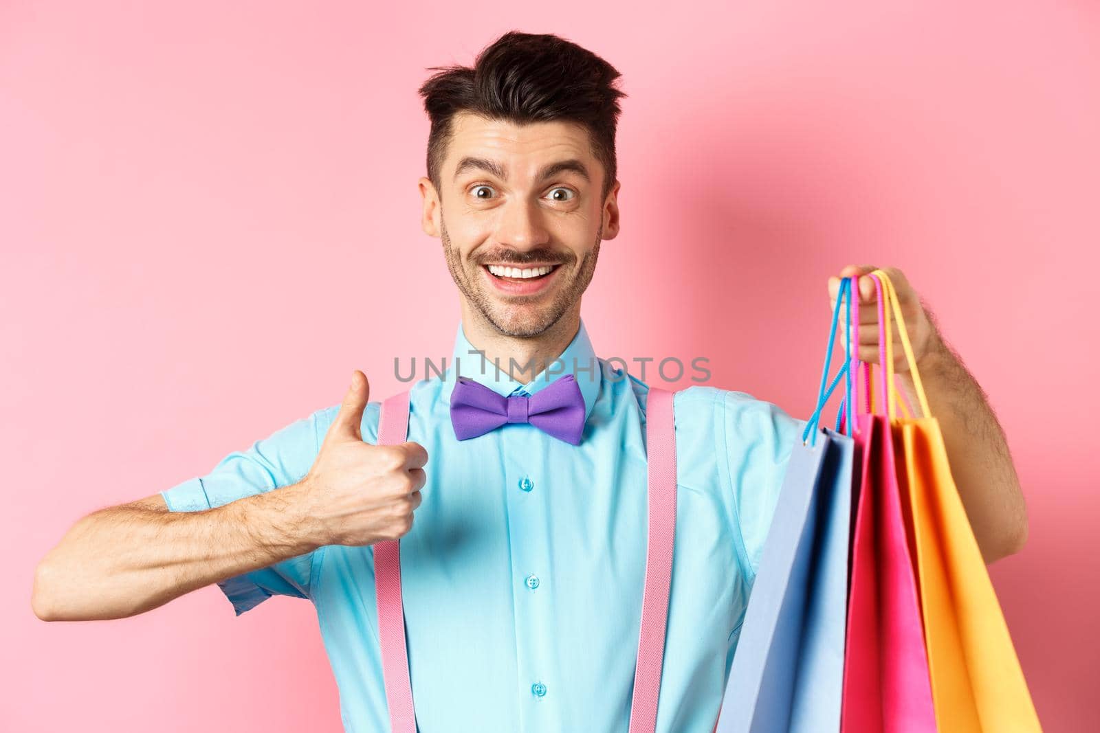 Happy male shopper showing thumbs up and shopping bags, recommending store, standing on pink background.