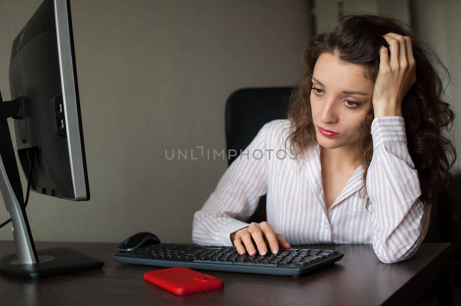 Tired young woman with curly hair and white shirt is working at the office using her laptop, routine work, freelance, burnout syndrome.
