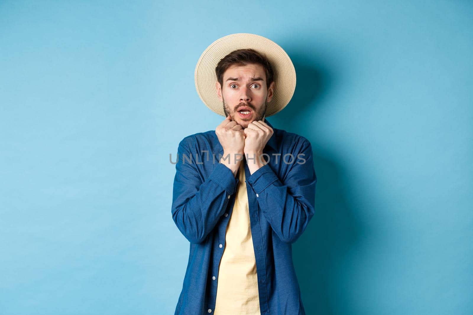 Scared young tourist trembling from fear, looking at something scary on summer vacation, wearing straw hat, standing on blue background by Benzoix