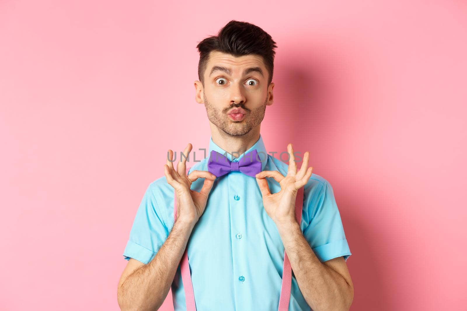 Handsome and funny young man pucker lips for kiss, showing his classy bow-tie, wearing clothes for party, standing on pink background.