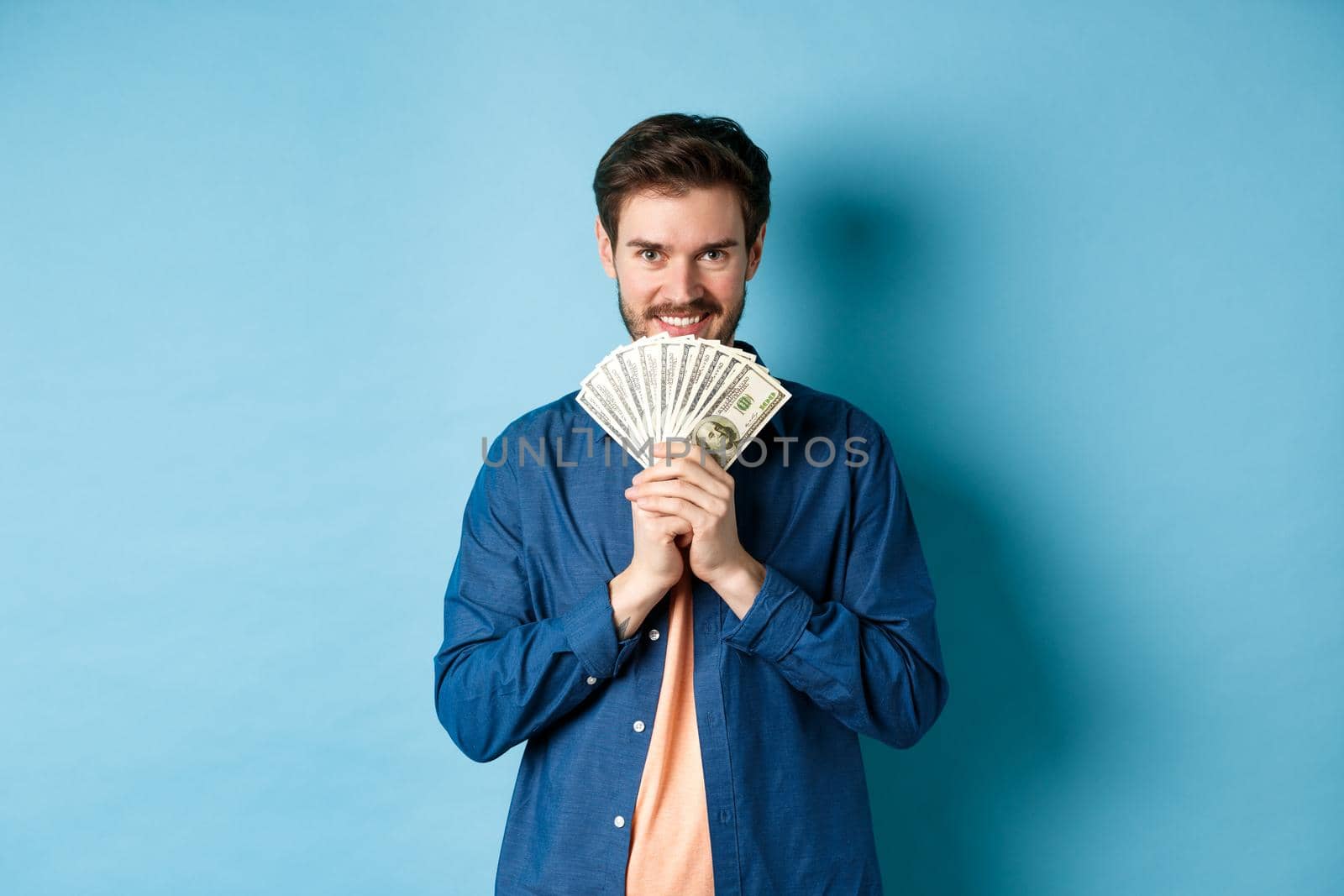 Happy smiling man holding money and looking at camera pensive, thinking of shopping, standing on blue background.