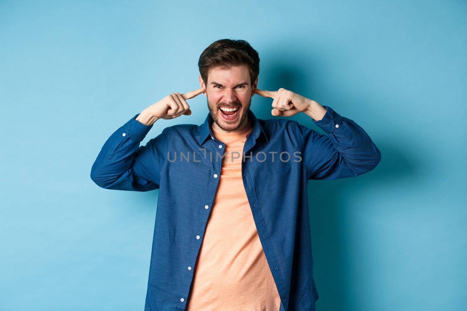 Angry young man shut ears to block annoying sound and shouting at camera, frowning and looking furious, standing on blue background by Benzoix