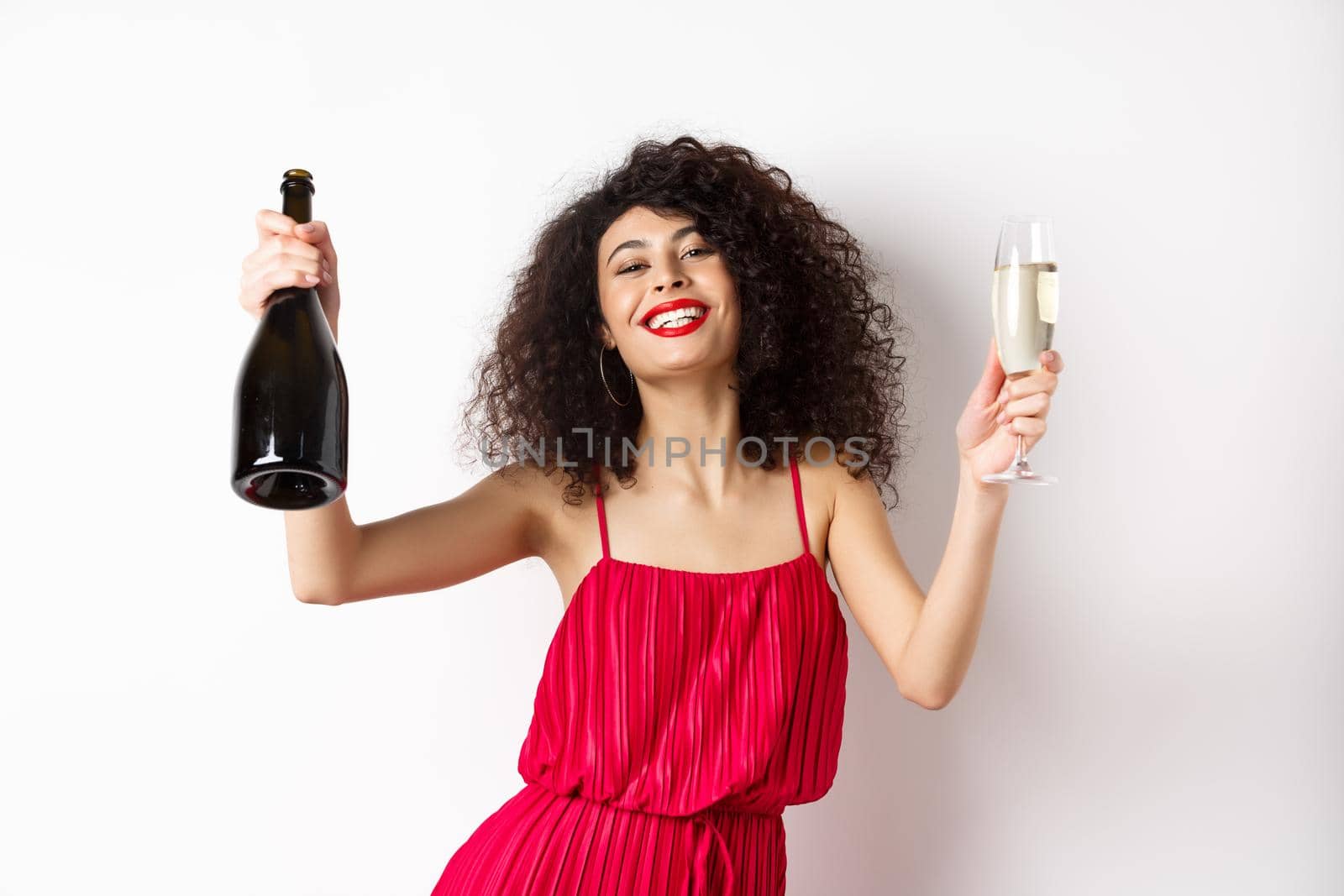 Happy party girl in red dress, dancing with bottle of champagne and glass, drinking and having fun, celebrating holiday, standing on white background.