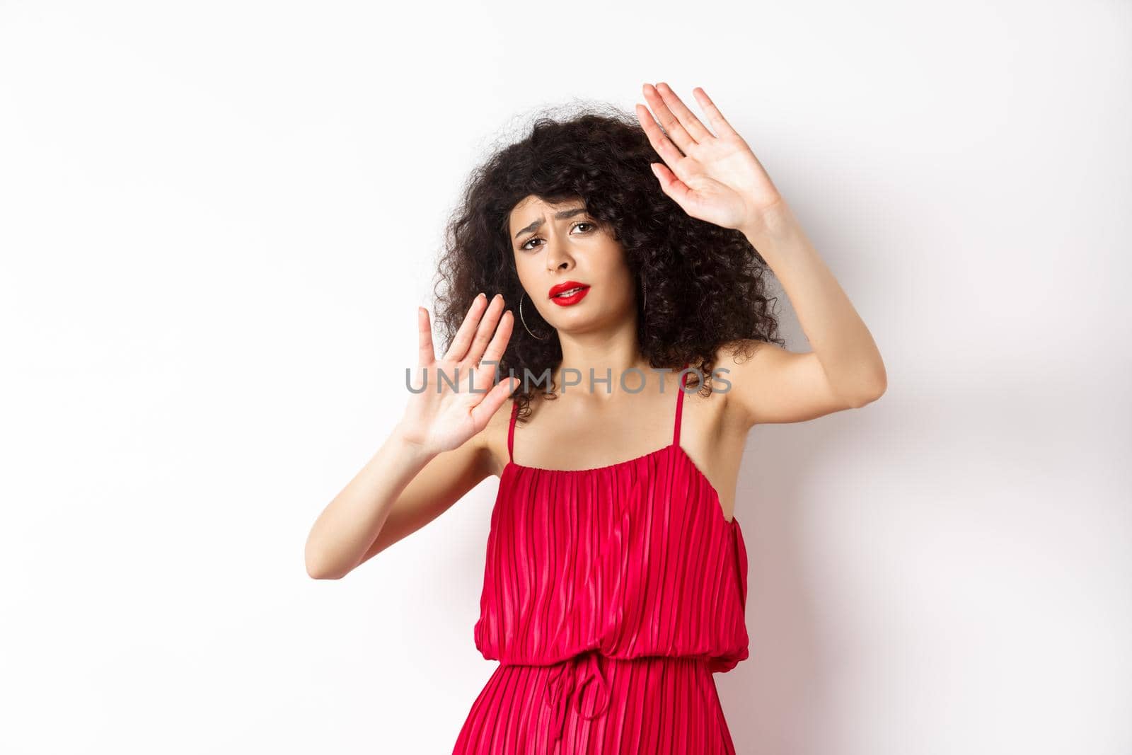 Young woman with curly hair and red dress, asking to stop, block someone, raising hands defensive, protecting herself, standing against white background.
