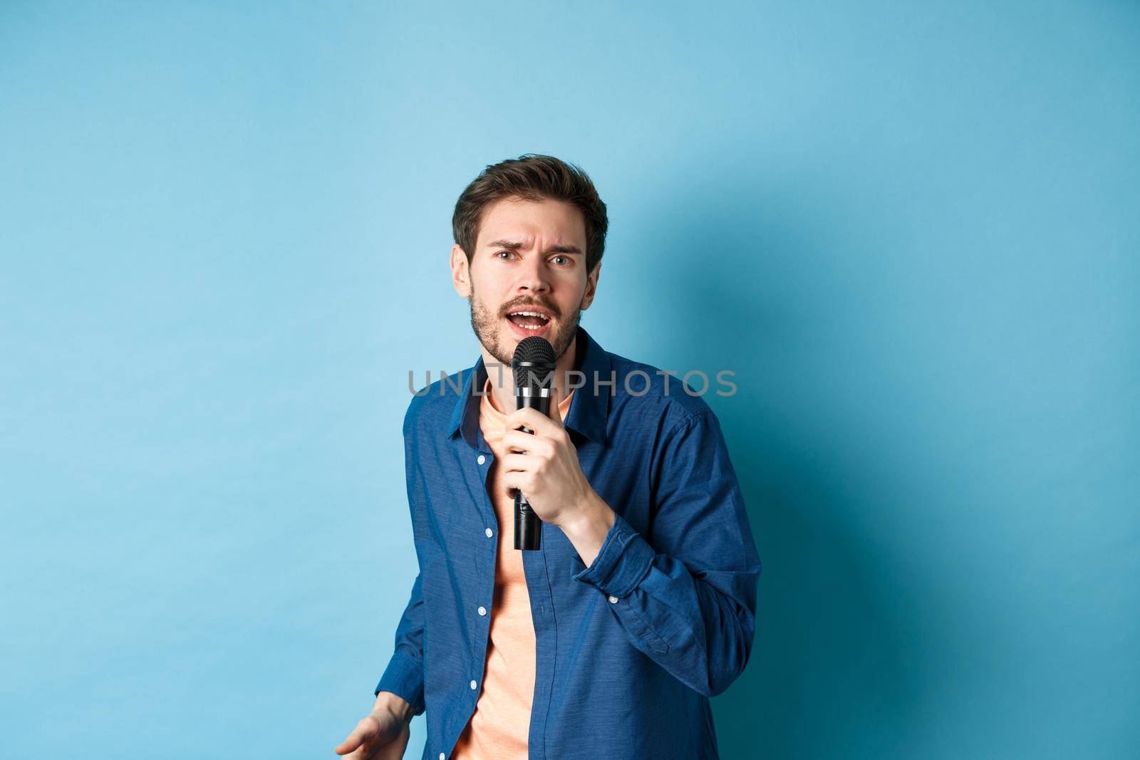 Passionate singer looking at camera, singing in microphone, playing karaoke on blue background. Copy space