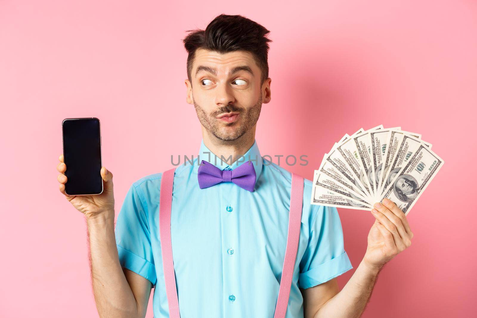 E-commerce and shopping concept. Funny guy look curious at empty smartphone screen, showing money, buying online, standing on pink background by Benzoix
