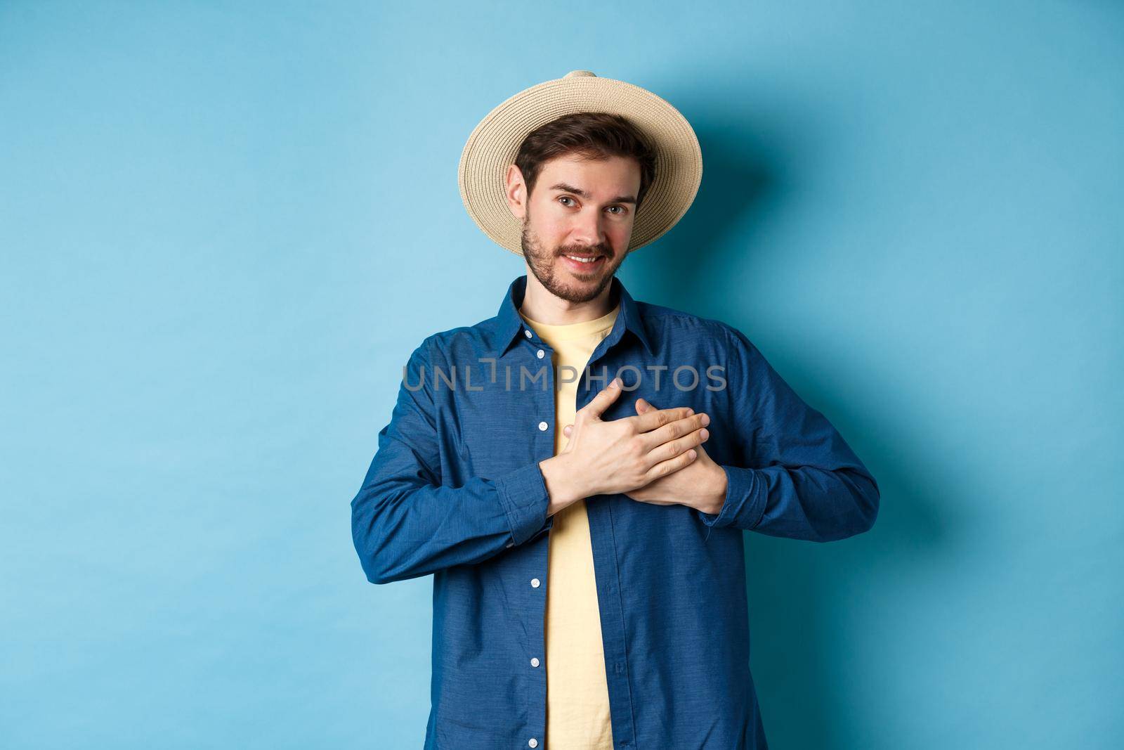 Handsome traveler in straw hat, holding hands on heart and smiling, showing thank you gesture, keep memories about vacation in soul, standing on blue background.
