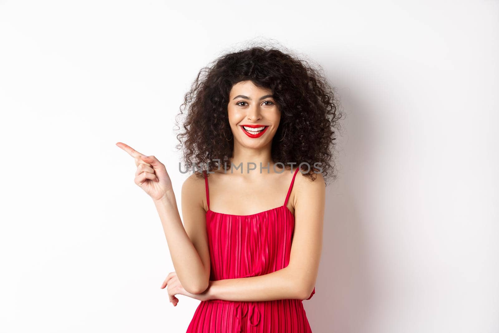 Beautiful happy woman with curly hairstyle, wearing festive dress, pointing finger left at logo and smiling, white background.