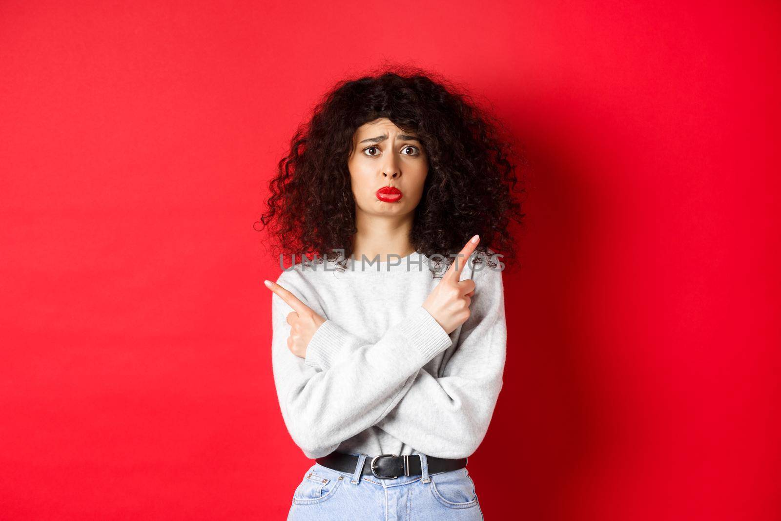 Nervous sad girl with curly hair, sobbing miserable and pointing fingers sideways, showing two things and look indecisive, red background.