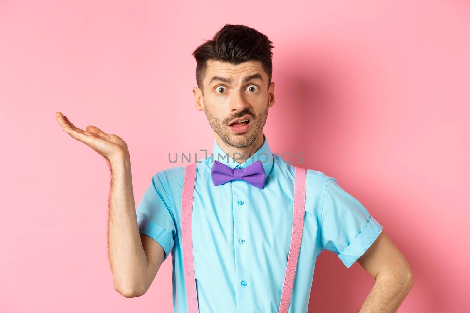 Confused guy cant understand what happening, looking with wtf face, raising hand and staring at camera shocked, standing over pink background.