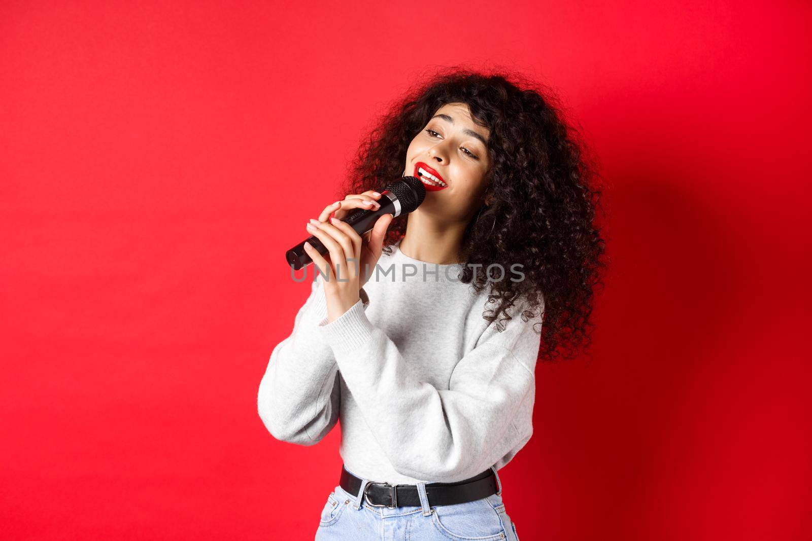 Leisure and hobbies concept. Stylish young woman singing karaoke, looking aside and holding microphone, performing song, standing on red background by Benzoix
