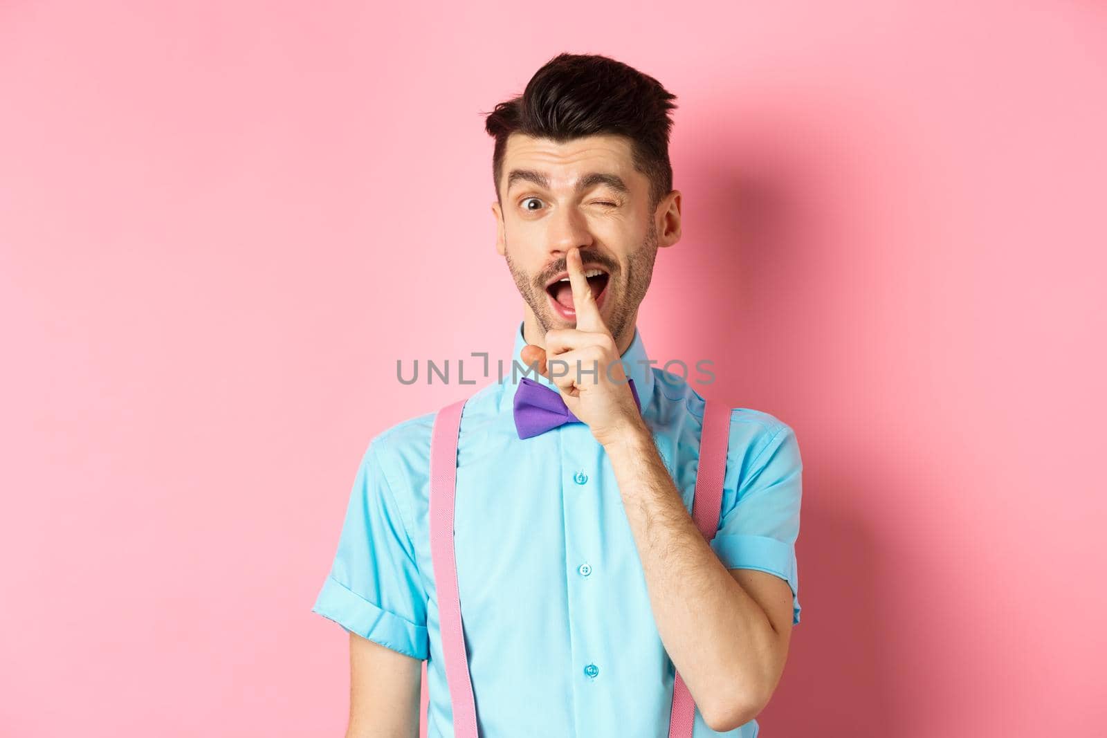 Funny young man planning a surprise, hushing and winking at camera, asking to keep quiet, telling a secret, standing on pink background by Benzoix