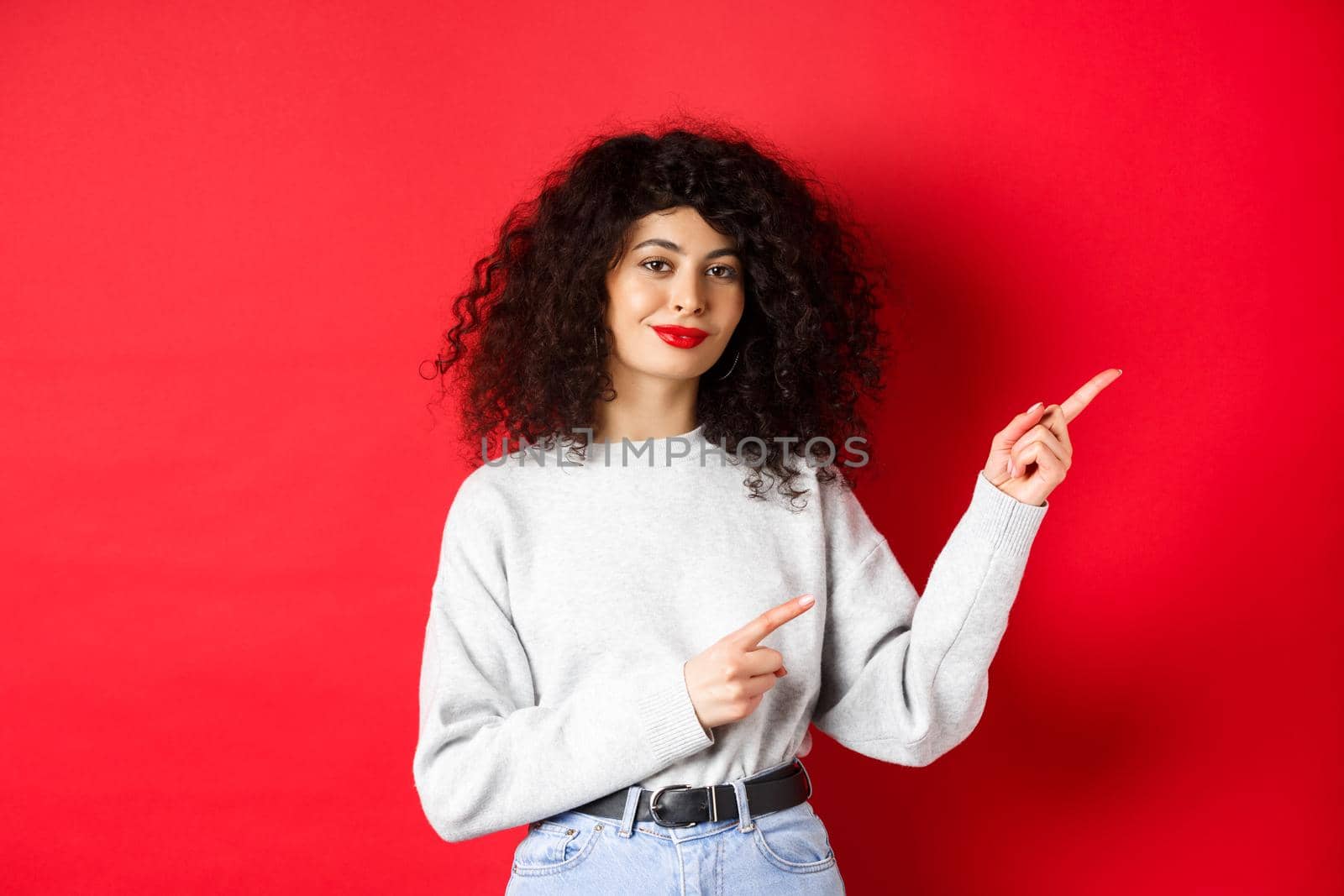 Smiling beautiful girl with curly hair, pointing fingers right at logo, showing advertisement, red background.