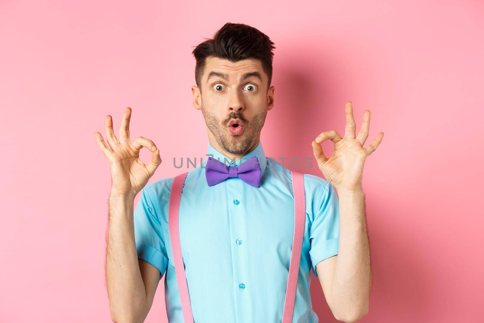 Impressed caucasian guy in bow tie, checking out cool offer, showing okay gesture and saying wow, praising good product, standing on pink background.
