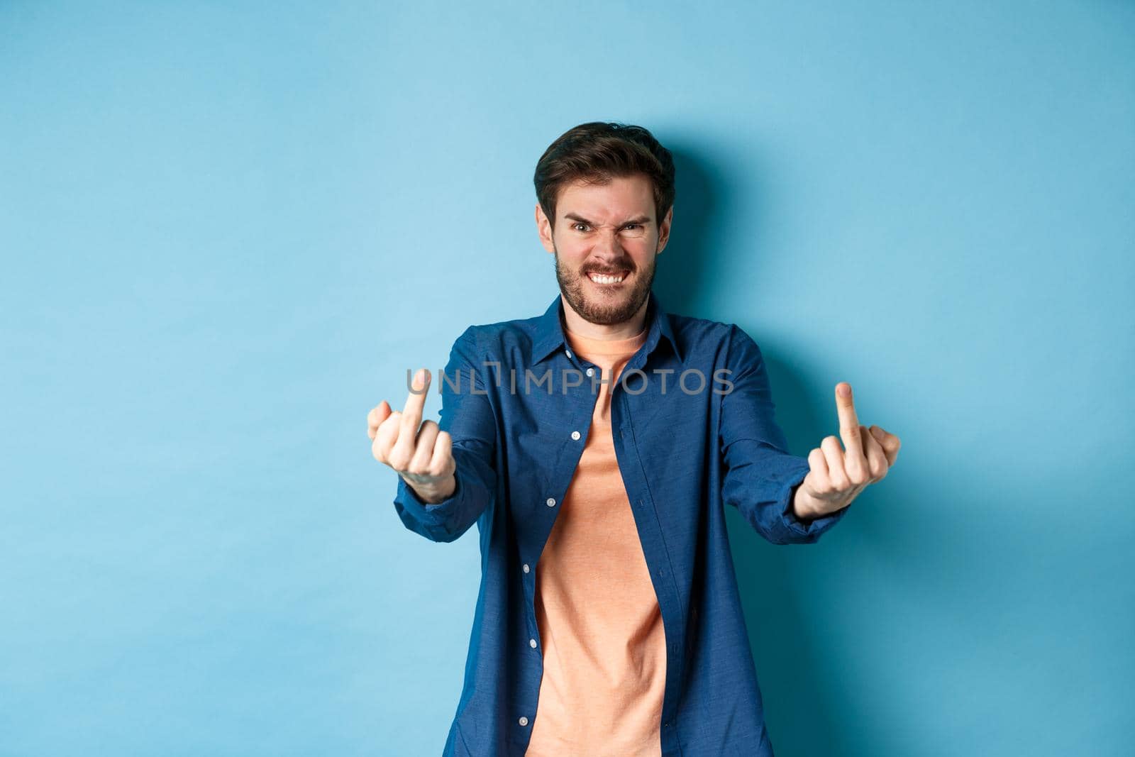 Angry caucasian man say fuck you and showing middle fingers, make rude gesture with pissed-off face, standing annoyed on blue background.