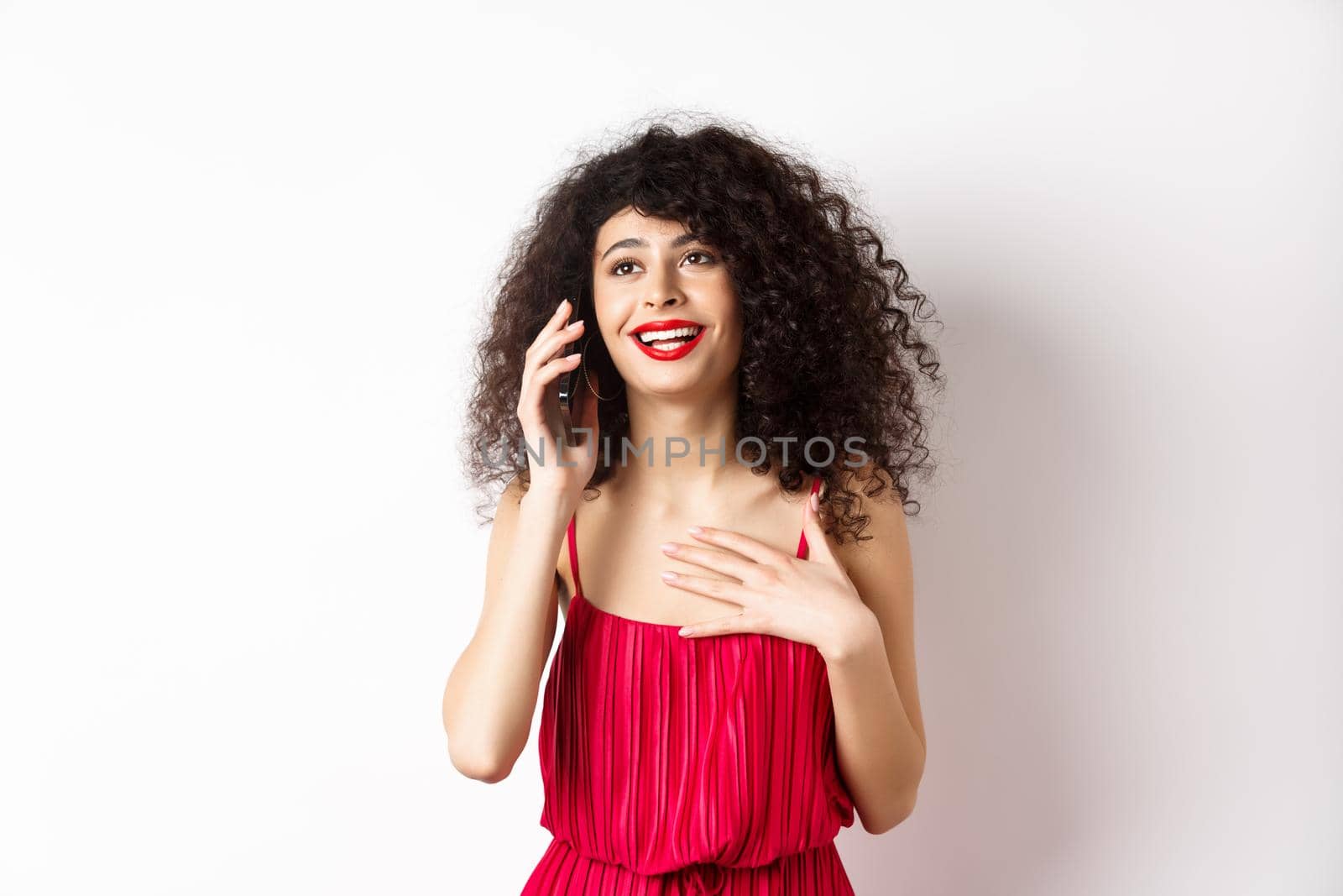 Beautiful lady with curly hair, wearing red dress and talking on phone, happy face, having pleasant conversation, standing on white background.