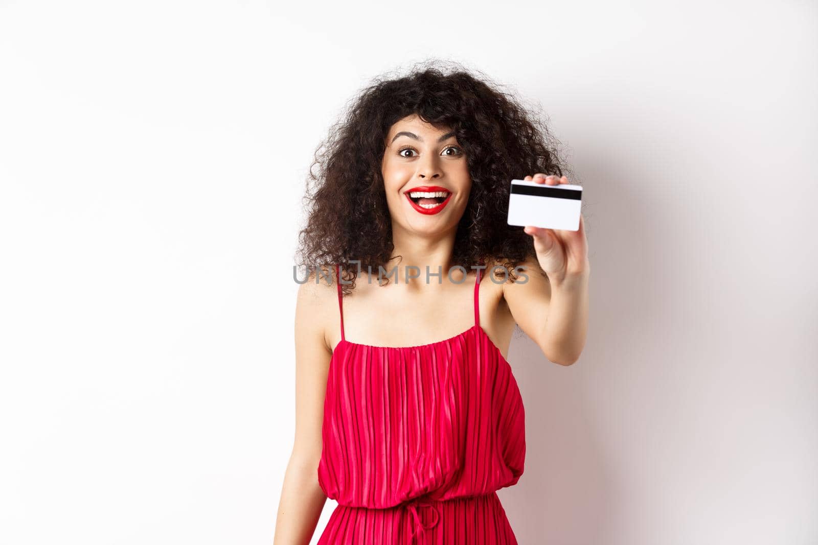 Shopping. Excited stylish lady with makeup, red dress, showing plastic credit card and smiling amazed, standing against white background.