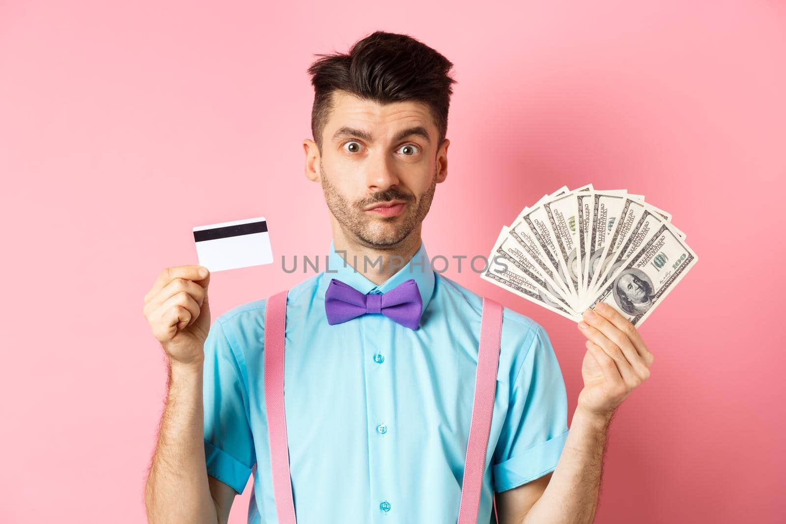 Handsome man in bow-tie showing plastic credit card and money in dollars, making an offer, standing over pink background. Copy space