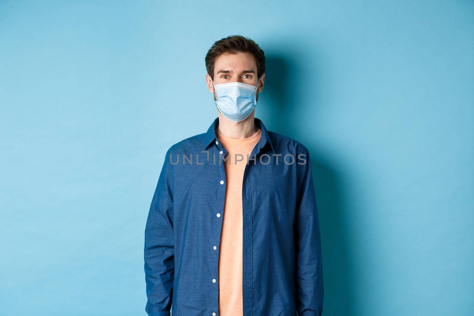 Covid-19 and healthcare concept. Smiling young man in medical mask looking healthy and happy, standing on blue background.