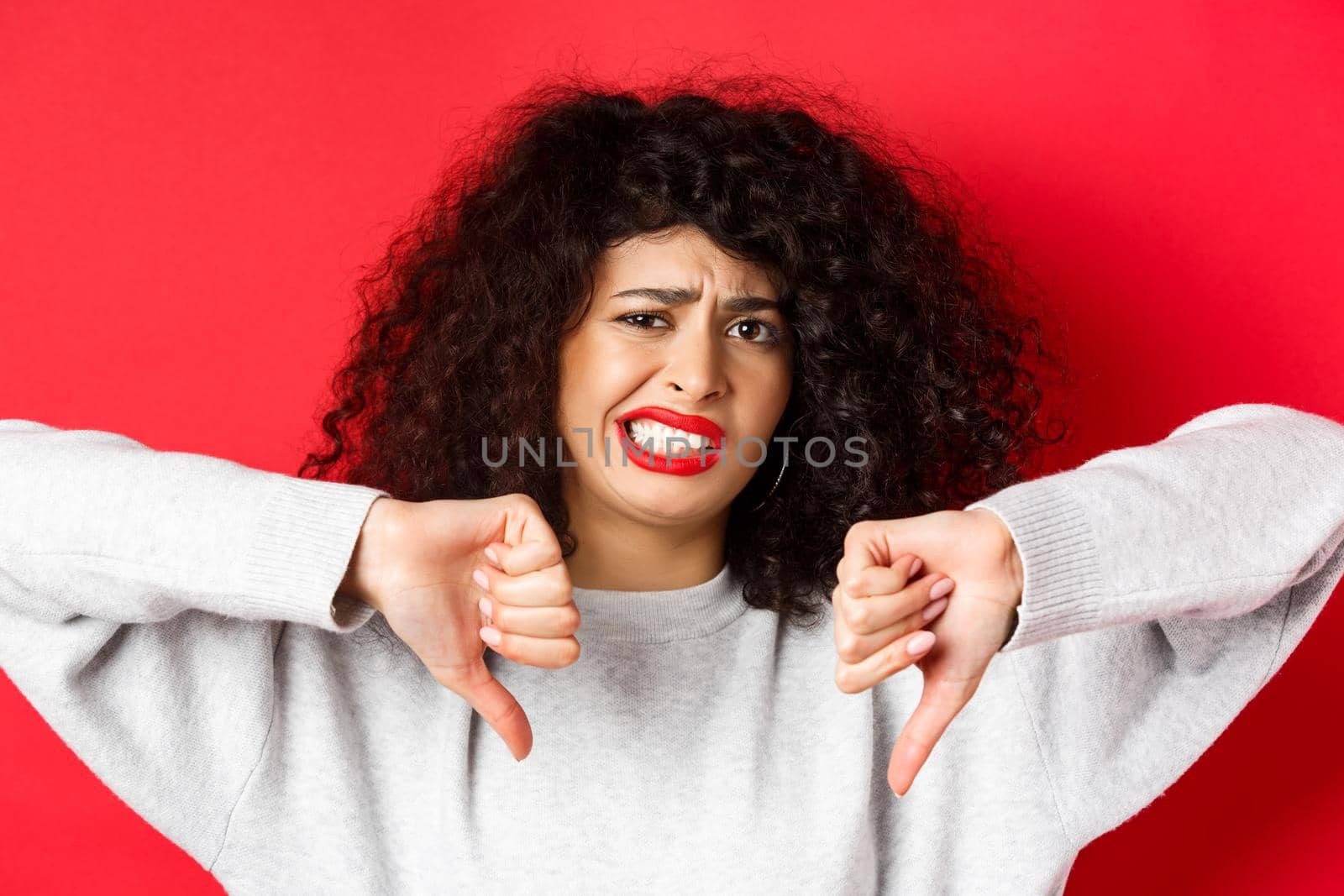 Close-up portrait of disappointed woman grimacing, cringe from something bad, showing thumbs-down, dislike and disapprove, standing on red background.