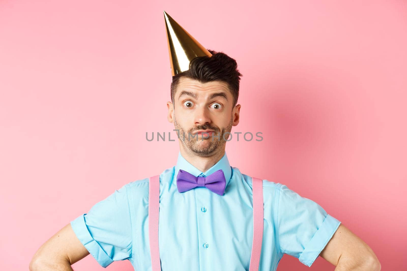 Holidays and celebration concept. Close-up of confused male entertainer in party hat and bow-tie, looking puzzled and shocked, standing over pink background.