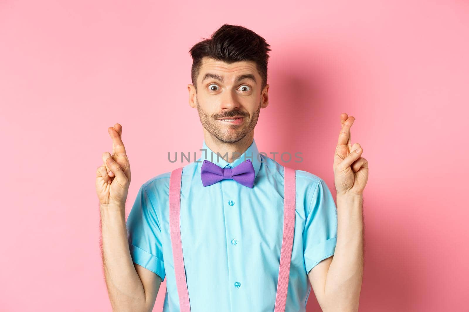 Hopeful young man making wish, holding fingers crossed and smiling, waiting for results with optimism, standing over pink background.