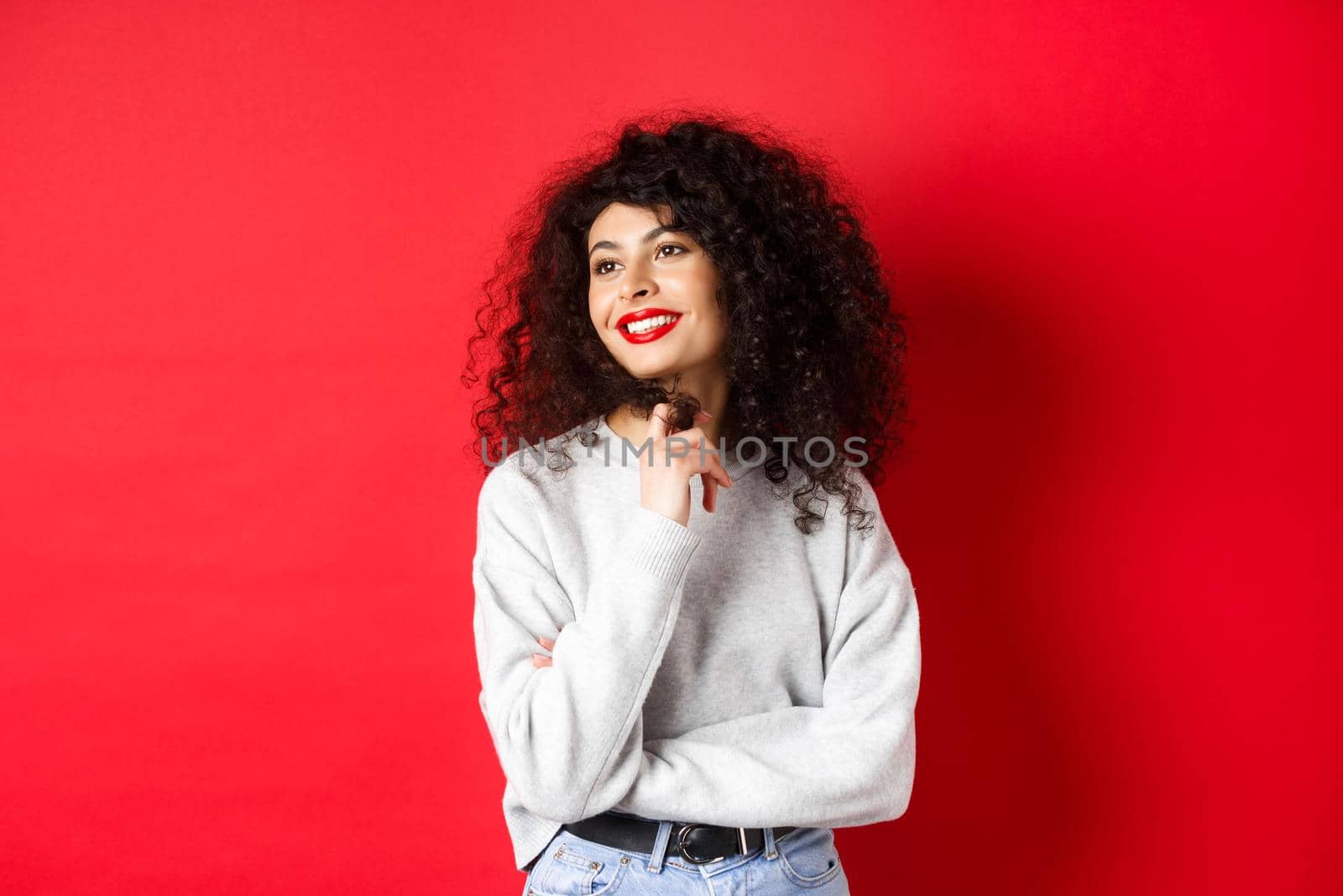 Beautiful young woman with curly hair and red lips, playing with curl and looking aside, gazing romantically, standing on studio background.