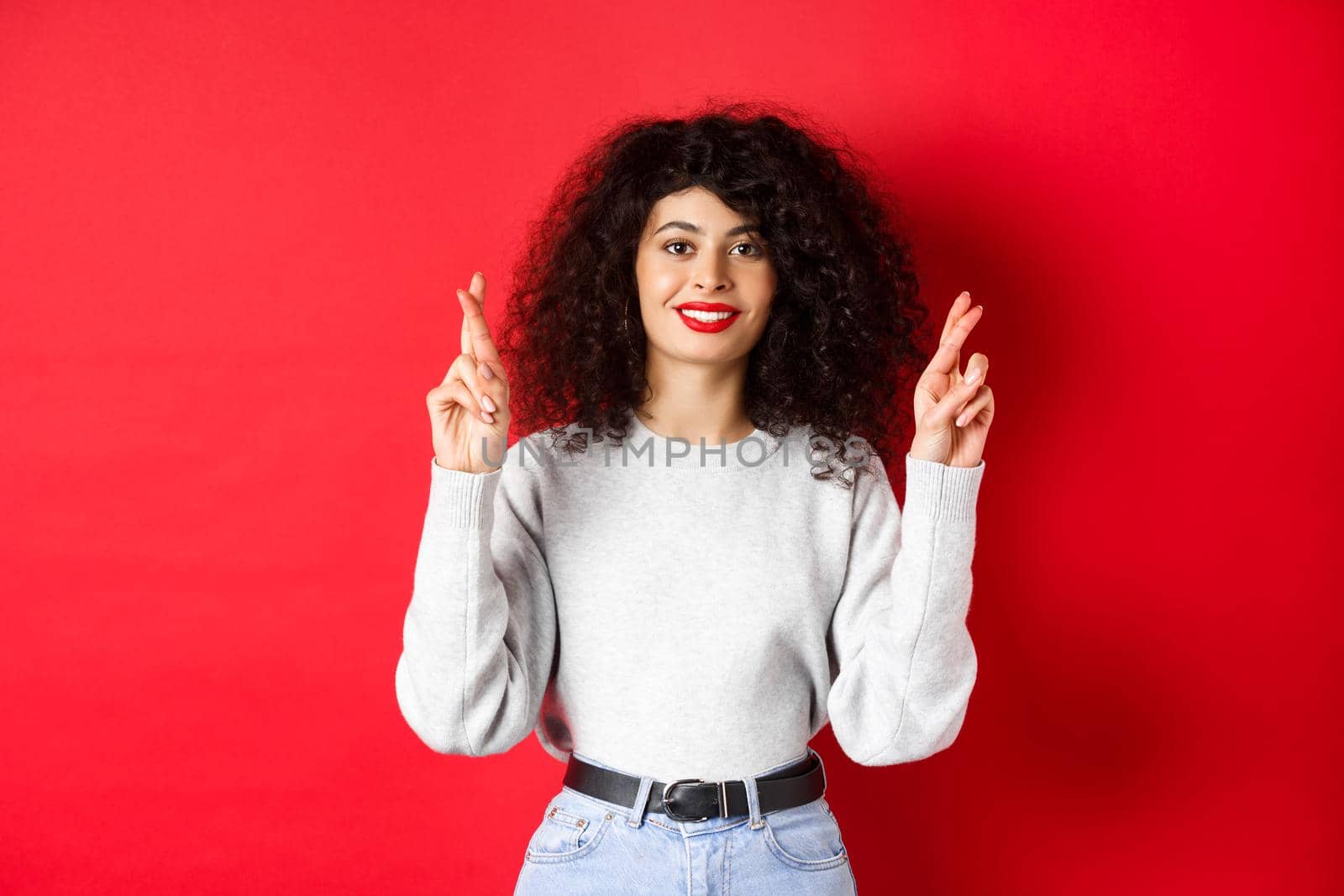 Hopeful young woman with red lips and curly hair, cross fingers for good luck and making wish, praying for dream come true, smiling excited, red background.