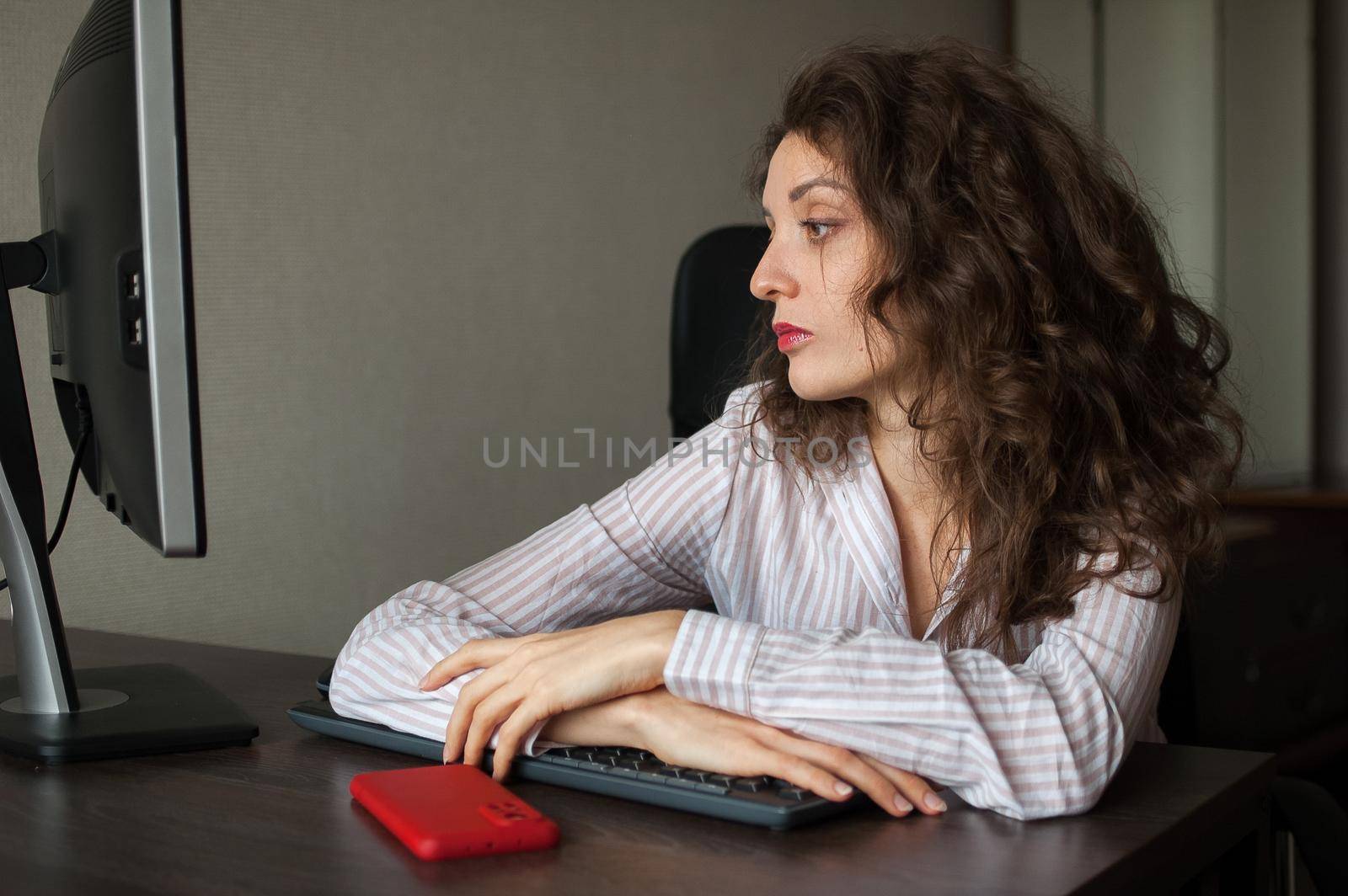 Tired young woman with curly hair and white shirt is working at the office using her laptop, routine work, freelance, burnout syndrome.