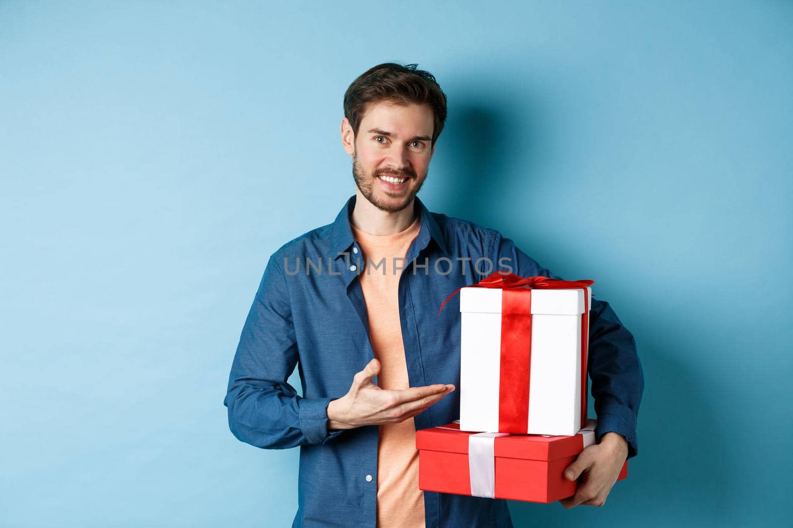Young man in casual clothes buying romantic gifts on Valentines day, pointing at presents boxes and smiling, standing over blue background.